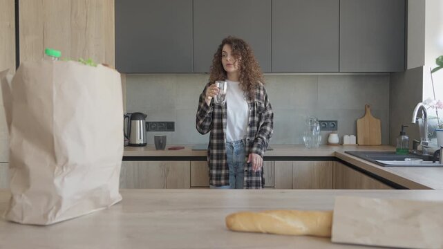 Woman Drinking Water After Grocery Shopping In Modern Kitchen
