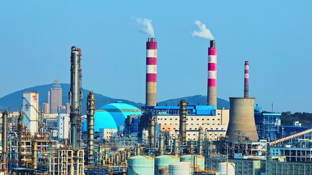 High angle view of an industrial refinery complex with tall red and white striped smokestacks emitting smoke under bright daylight and clear blue sky.