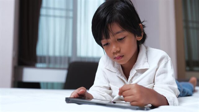 Asian boy drawing with stylus pen on digital tablet while lying on bed. Early childhood education, digital art, screen time, e-learning, and smart kid concept.