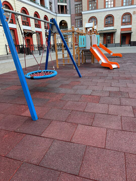 A colorful playground scene featuring a blue swing and orange slide set against a background of patterned red pavement. The setting is urban with buildings in view.