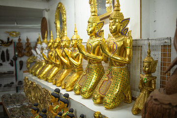 Fototapeta premium Large golden Thepphanom statues stand together with palms pressed in wai prayer pose, traditional Thai guardian figures display intricate craftsmanship inside decorative art shop