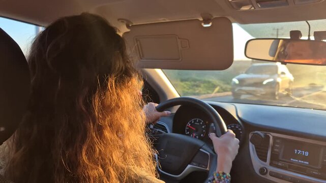 Woman driving car during golden hour sunset on highway, holding wheel with sunset light, medium static interior camera shot.