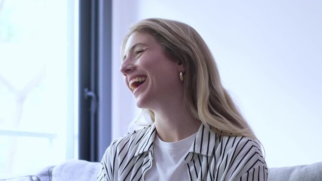 Close-up of a happy young woman with blonde hair laughing genuinely while sitting on a comfortable sofa at home, her expression transitioning from pure joy to a calm, relaxed state