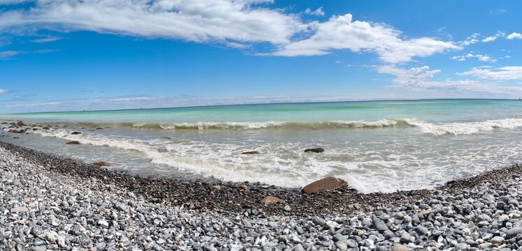 Wide curved perspective of waves crashing onto dark pebble shoreline 