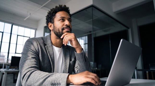 Pensive Black male executive with beard sitting in a modern corporate office behind laptop, hand under his chin, focused on long term financial planning and strategic business management.