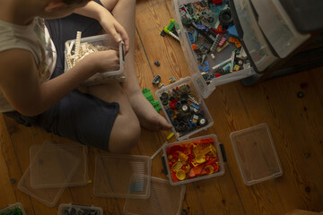 Obraz premium overhead view of child sitting crosslegged on wooden floor sorting multiple clear boxes of colorful plastic building bricks and wheels with hands focused on white