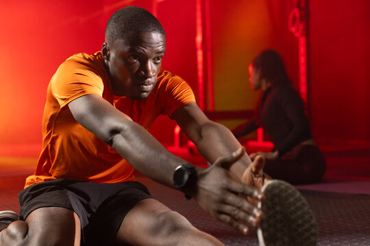 African American partners stretching on gym floor under red lights, orange shirt and smartwatch