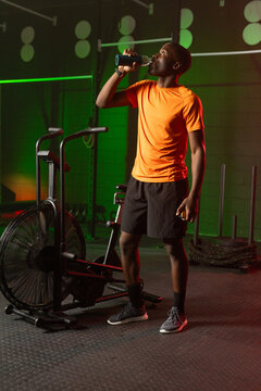 African American man in orange tee drinking from bottle holding fruit by air bike in gym