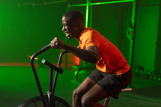 African American man pedaling intensely on air bike at gym, wearing orange shirt, copy space