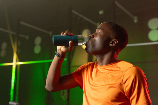 African American adult man pausing workout, drinking from reusable bottle in gym with orange shirt