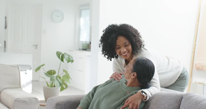 African American adult approaching behind, leaning over senior woman on gray sofa at home