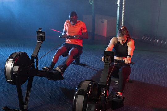 Workout partners rowing on machines in dim studio featuring flywheels, one in orange