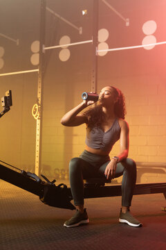 African American woman sitting on rowing machine drinking from reusable bottle in gym