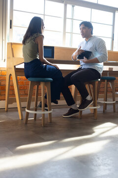 Colleagues discussing work on laptop and tablet seated on stools at wooden counter beneath windows