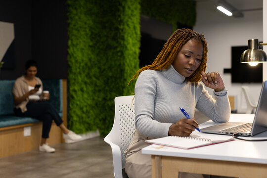 African woman working on laptop and writing notes at coworking lounge with desk lamp