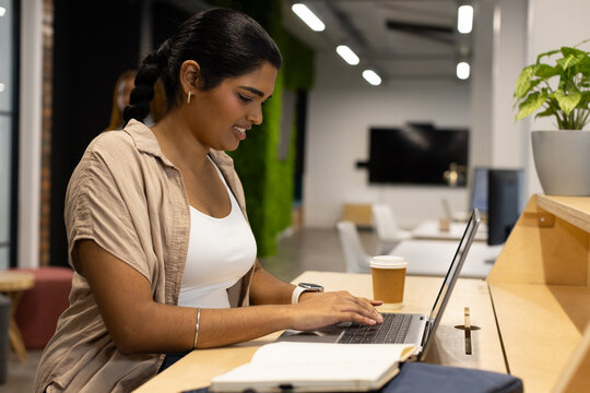 Indian woman working on laptop at high wooden counter in coworking lounge, wearing beige overshirt