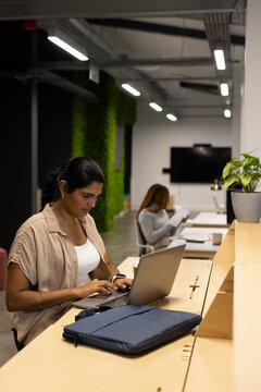 Diverse female colleagues typing and reading at co-working wooden counter with laptop papers plants