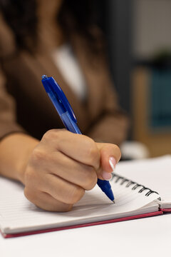 Female in brown blazer holding blue ballpoint pen poised over spiral notebook on white table