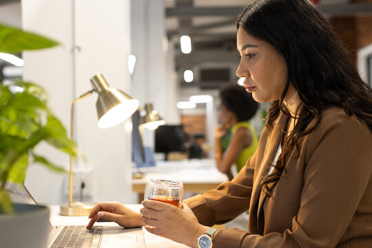 Diverse female coworkers working in open-plan office with laptop, glass and brass lamp, copy space