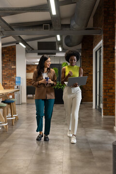Diverse female coworkers wearing blazers walking and discussing in office holding laptop