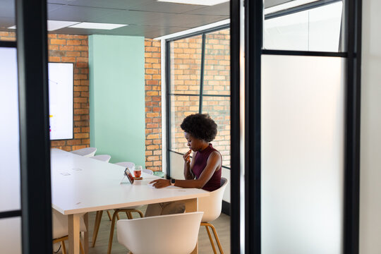 African woman in burgundy top sitting at table in room, snacking while reviewing tablet, copy space