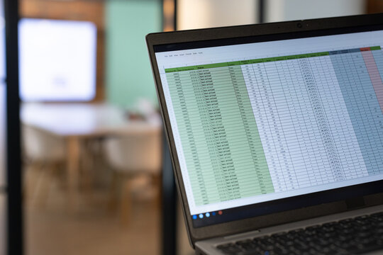 Laptop showing spreadsheet with green column in right foreground, conference room blurring