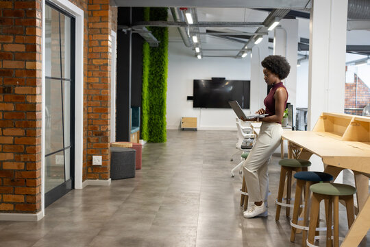 Woman in 20s standing at wooden counter using laptop near moss panel in office, copy space