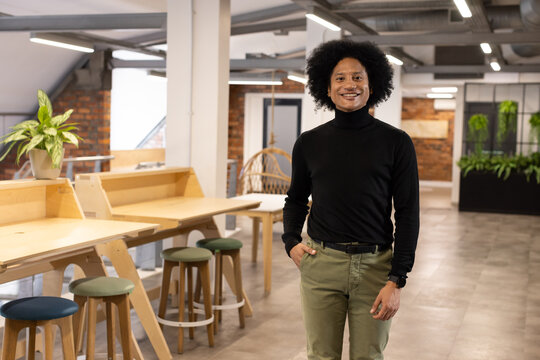 Adult man wearing black top standing in coworking office by desks and wicker chair, checking watch