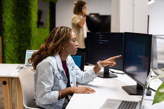 Diverse coworkers at desk with woman pointing at dual monitors while man talking on phone