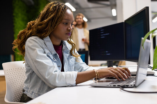 Coworkers collaborating, woman wearing denim jacket typing on laptop at office with code on monitor