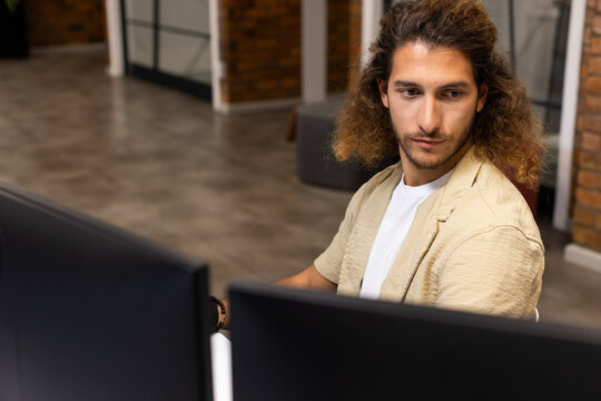 Man with curly hair wearing beige blazer working at desk with dual monitors, bracelet, copy space