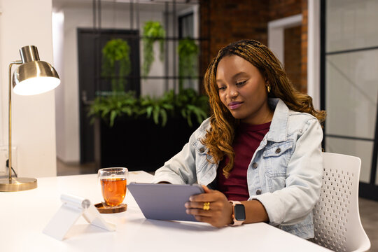 African American woman wearing denim jacket sitting at desk in office using tablet, brass lamp, tea