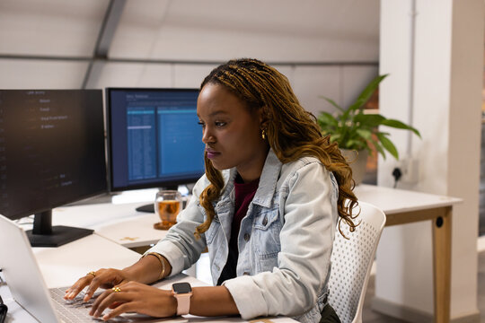 African American woman typing on laptop in denim jacket at office desk with dual monitors, mug