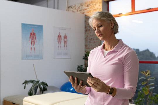 Senior woman standing in clinic by treatment table in pink polo, holding tablet reviewing charts