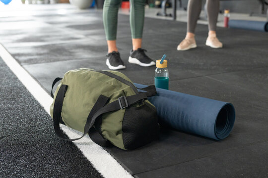 Women workout partners standing by green duffel, blue mat and bottle at gym wearing leggings, shoes