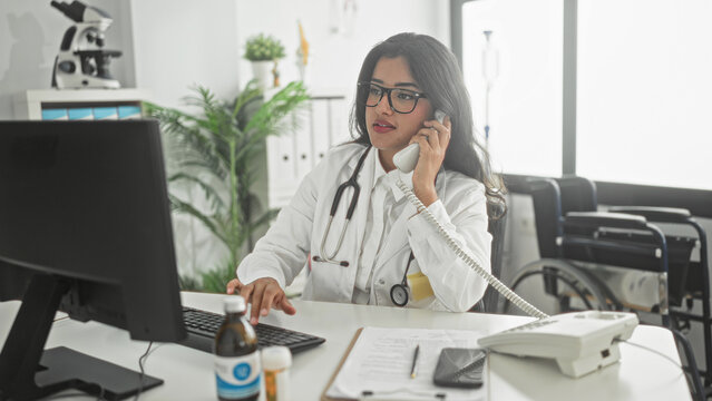 Woman doctor holding phone handset and typing on computer at desk in clinic building; focused care coordination and triage.