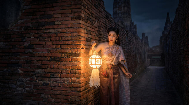 Thai woman in traditional costume holding glowing paper lantern walking through ancient brick temple ruins