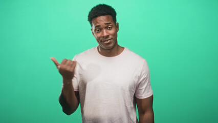 Fototapeta premium Man pointing thumb to side in studio with green backdrop, wearing pale pink tshirt and smirking with a shrug; playful.