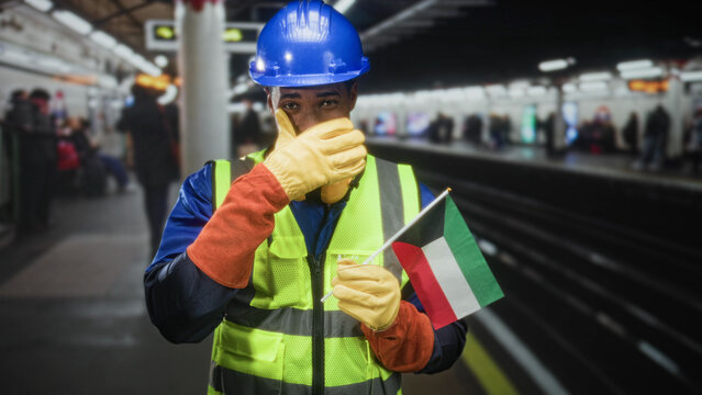 Man wearing blue hardhat and yellow vest holding kuwait flag and covering nose with gloved hand in building; safety concern.