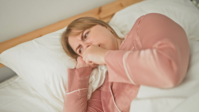 Woman coughing with hand to mouth on bed in bedroom, grimacing while lying on pillow; discomfort illness concern.