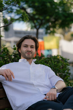 A handsome young man in a white shirt is sitting on a bench and looking at the camera.