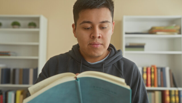 Man holding open book and reading intently in a library building with shelves behind, eyes on pages; quiet study calm.