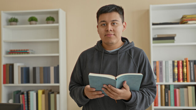 Man holding book in a library building, hands visible, wearing dark hoodie and standing by bookshelves while reading and gazing to the side; calm reflection.