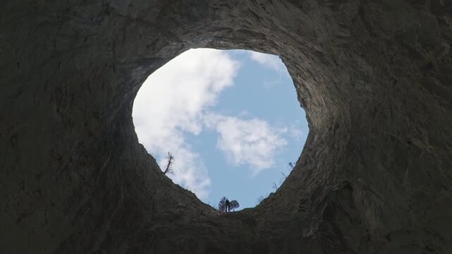 Dramatic low angle shot of an adventurer beginning to rappel down from the edge of the massive Coșul Boului rock portal in Romania, silhouetted against a blue cloudy sky.