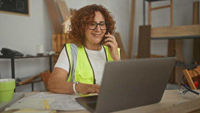 Woman carpenter holds phone to ear and types on laptop amid wood boards and safety glasses in a studio; focused planning.