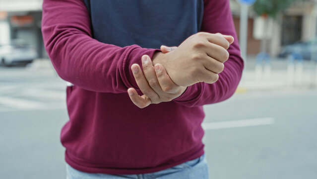 Young hispanic man clutching and rubbing his wrist on a city street crosswalk, wearing maroon pullover and jeans with hands central to frame; pain recovery.