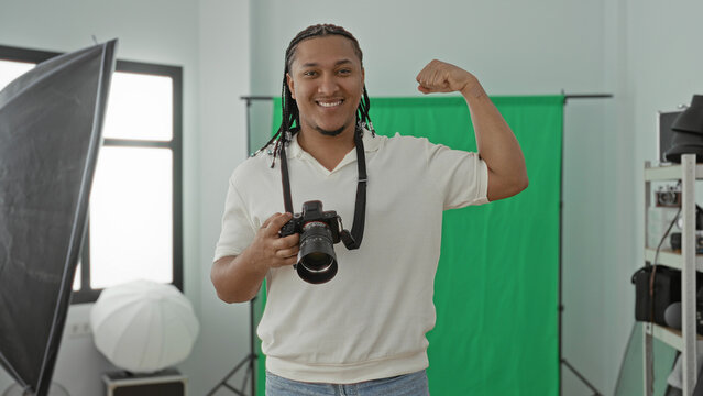Man photographer holds dslr camera and flexes arm in studio with green screen, softbox and window light; confidence success.