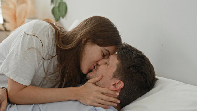 Man lies on bed pillow looking up as woman leans over smiling and touches his shoulder and face in a bedroom inside a building; affection warmth.