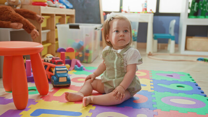 Obraz premium Toddler girl sitting on foam alphabet mat with bare feet and toy truck, looking up in a bright playroom; curiosity innocence.