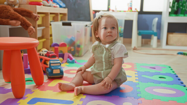 Toddler girl sitting on foam alphabet mat with bare feet and toy truck, looking up in a bright playroom; curiosity innocence.